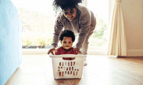 Mom pushing child around living room in laundry basket while child laughs and smiles